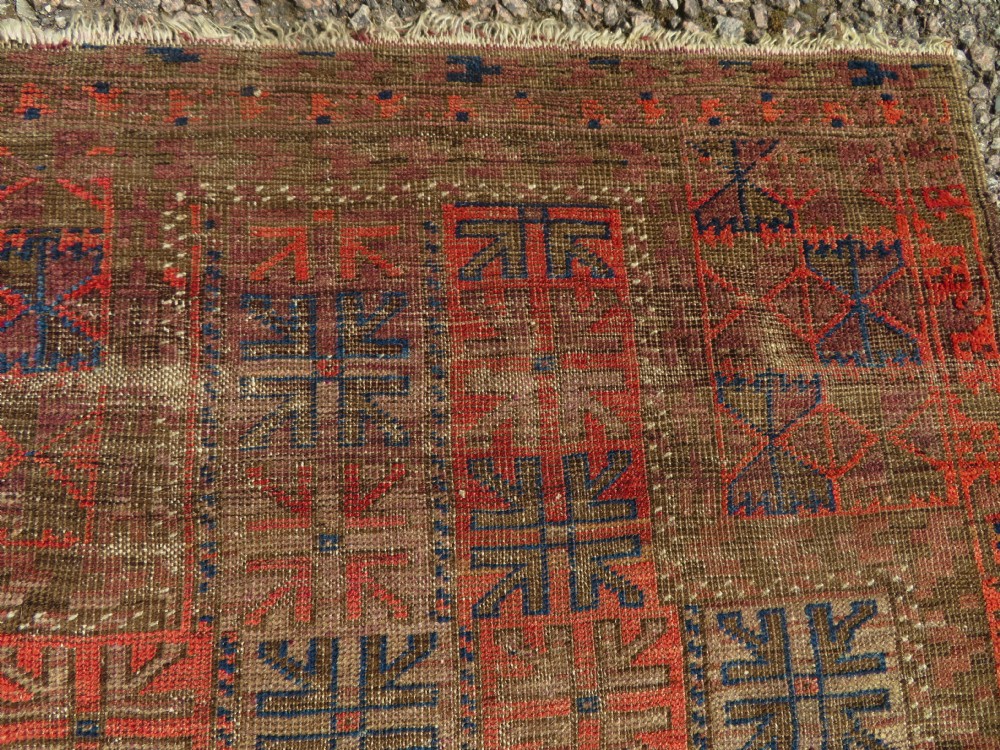 An antique hand-woven Baluch prayer rug showing distressed pile wear with geometric motifs in terracotta red, indigo, and deep brown. View III