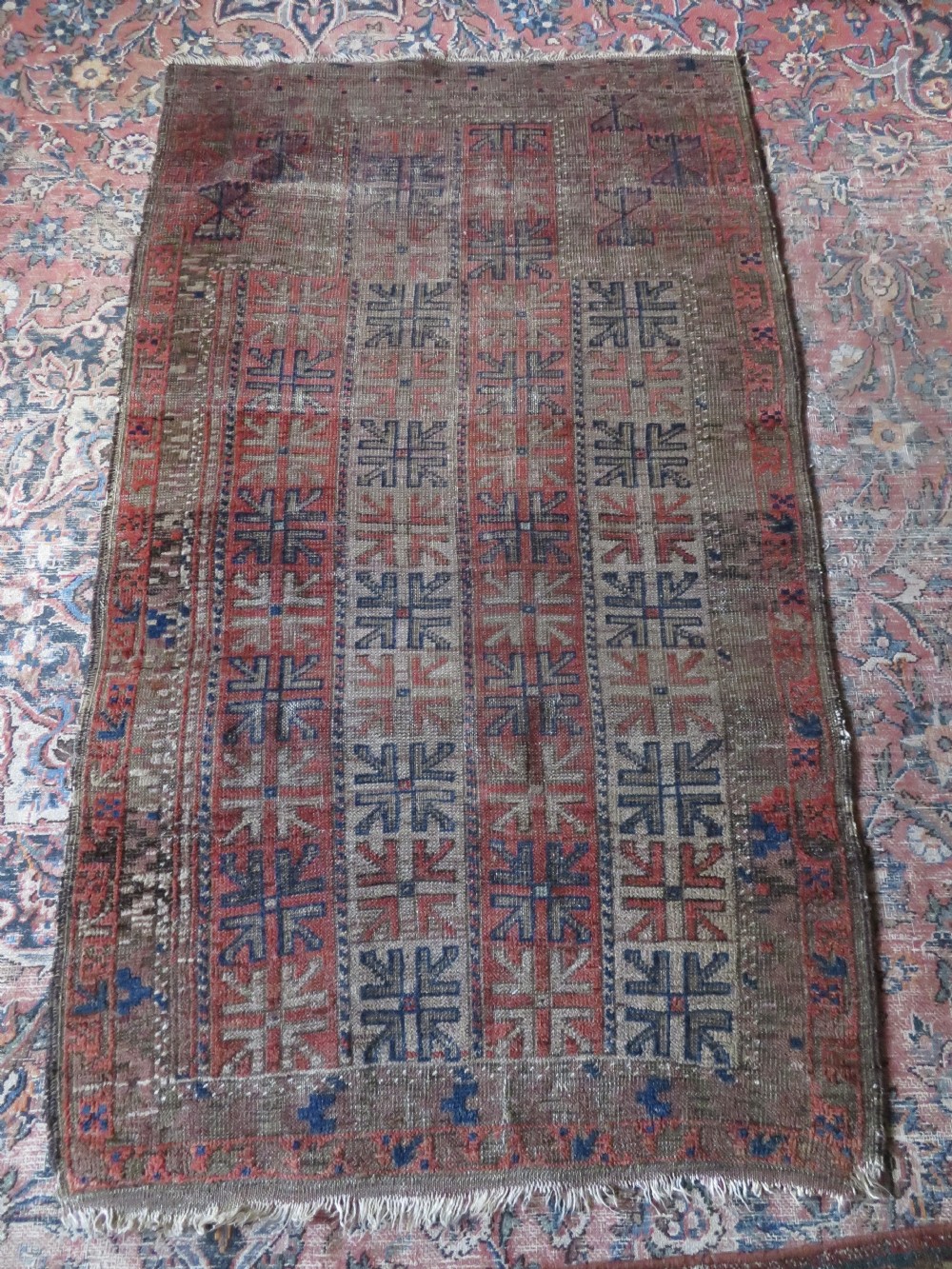 An antique hand-woven Baluch prayer rug showing distressed pile wear with geometric motifs in terracotta red, indigo, and deep brown. View X