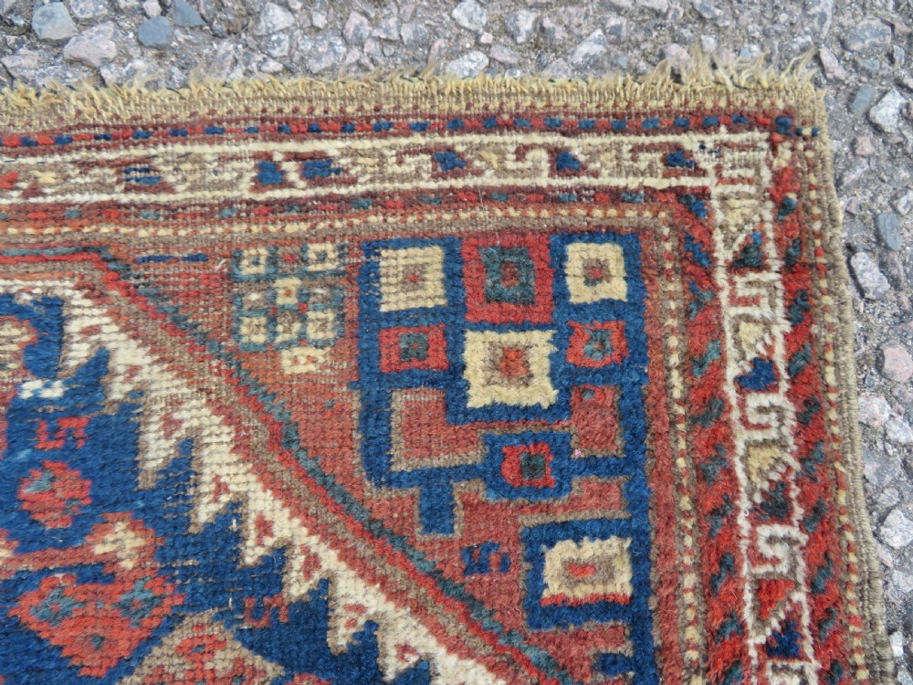 A small rectangular hand-knotted antique Khamseh rug featuring geometric tribal medallions in terracotta red, navy blue, and cream wool. View V
