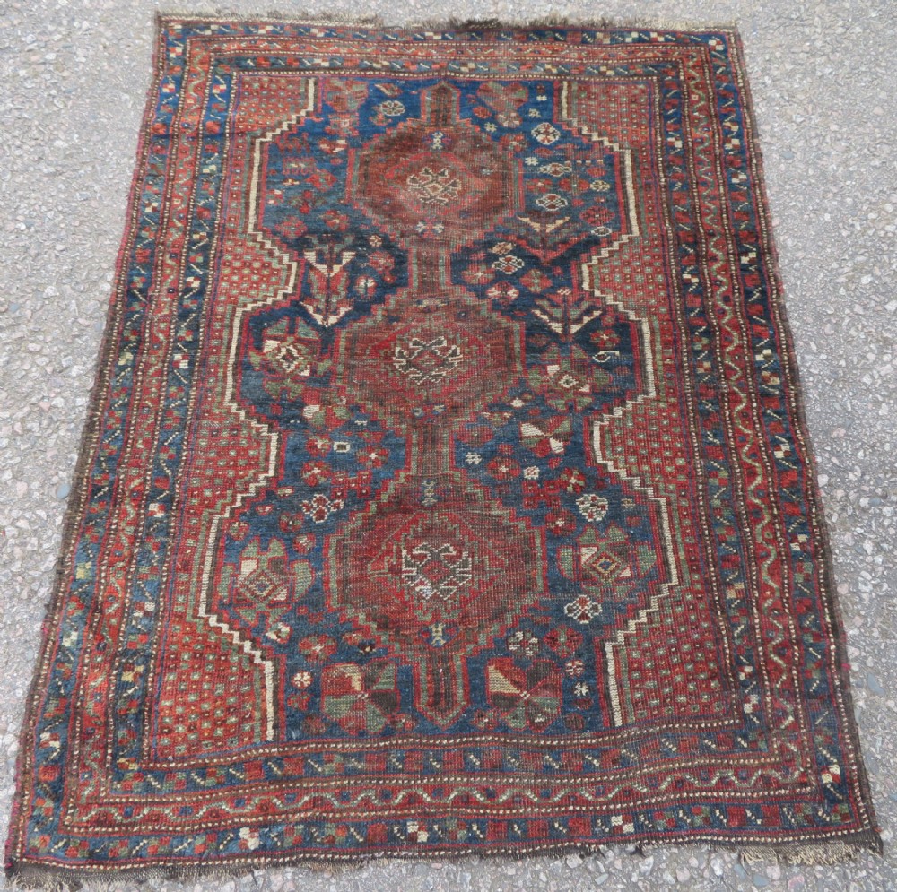 A distressed antique hand-knotted Khamseh tribal wool rug featuring three connected central medallions in red and indigo blue with geometric motifs.