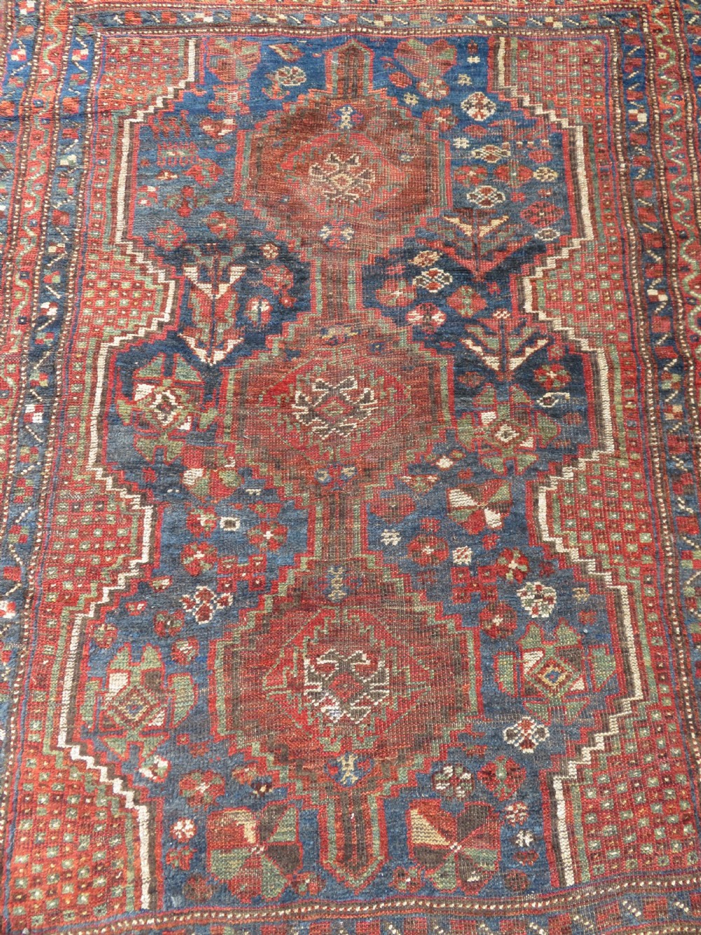 A distressed antique hand-knotted Khamseh tribal wool rug featuring three connected central medallions in red and indigo blue with geometric motifs. View III