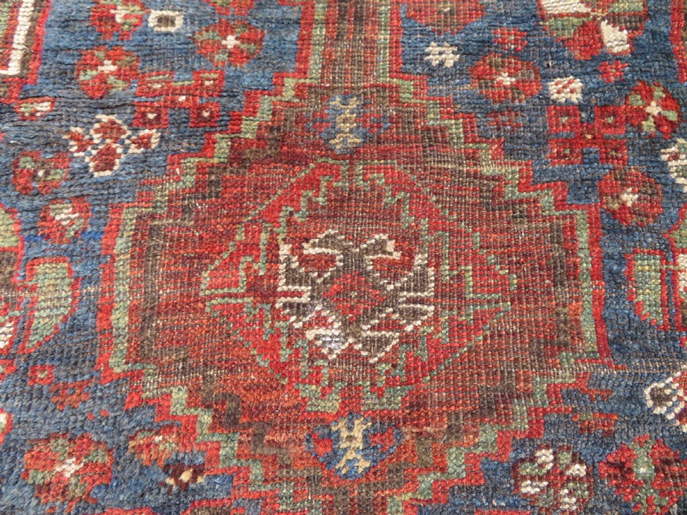 A distressed antique hand-knotted Khamseh tribal wool rug featuring three connected central medallions in red and indigo blue with geometric motifs. View V