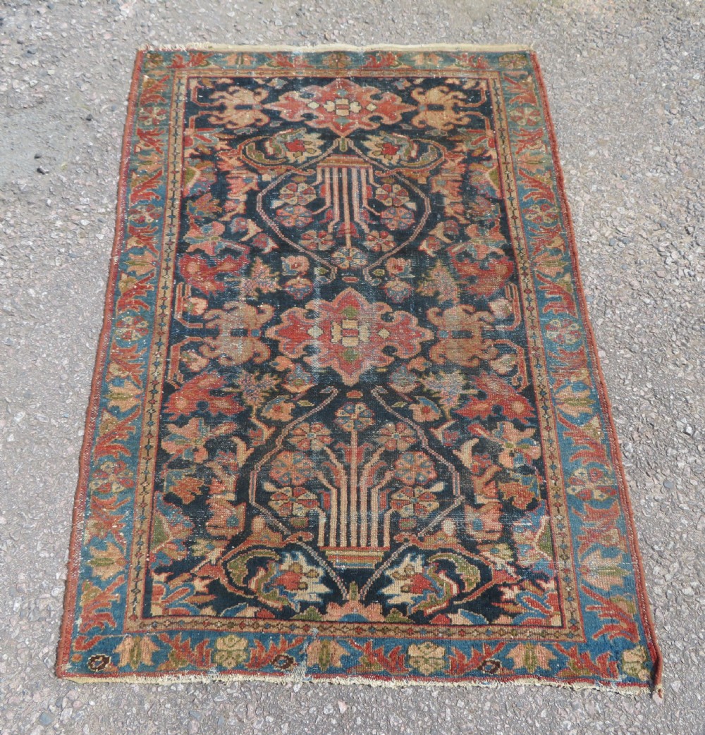 A small hand-knotted antique Sarouk wool rug featuring a dark blue field with central floral medallions and a terracotta and blue border.