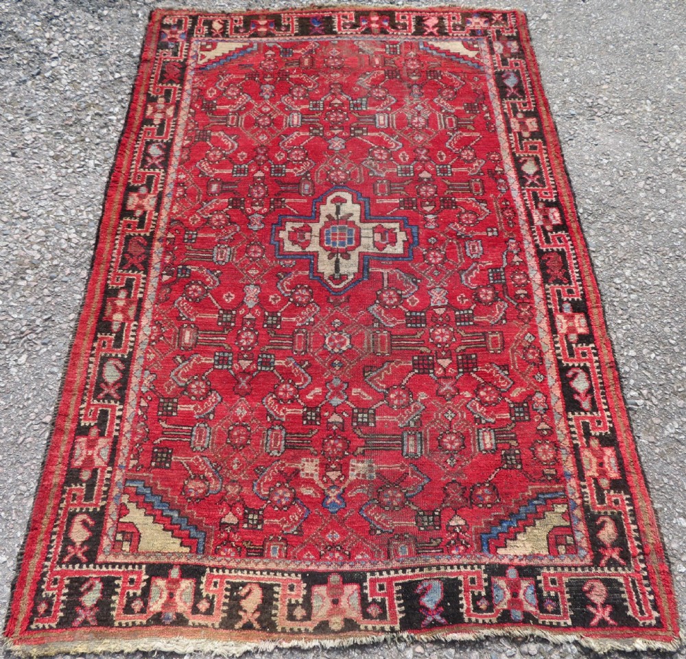 A rectangular distressed Persian Hamadan wool rug featuring a central stepped cross medallion on a crimson field with geometric borders.