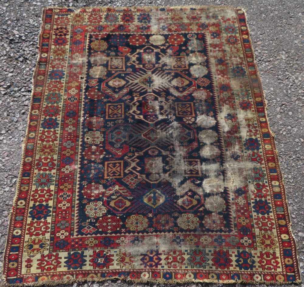 A rectangular hand-knotted antique Caucasian Daghestan rug featuring geometric medallions, intricate borders, and a distressed surface in red and navy blue tones.