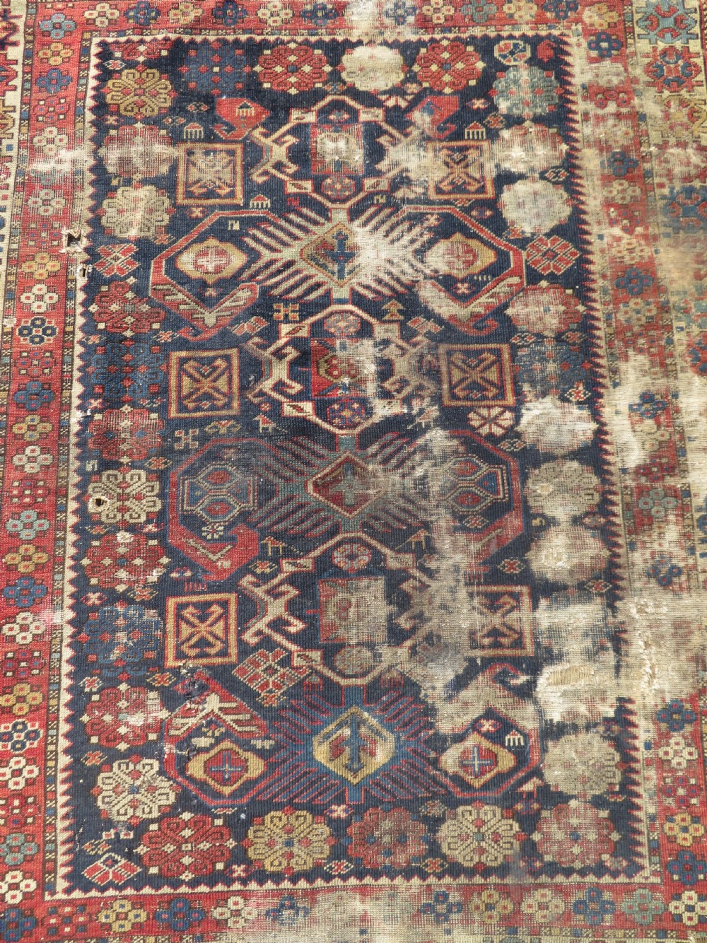 A rectangular hand-knotted antique Caucasian Daghestan rug featuring geometric medallions, intricate borders, and a distressed surface in red and navy blue tones. View II
