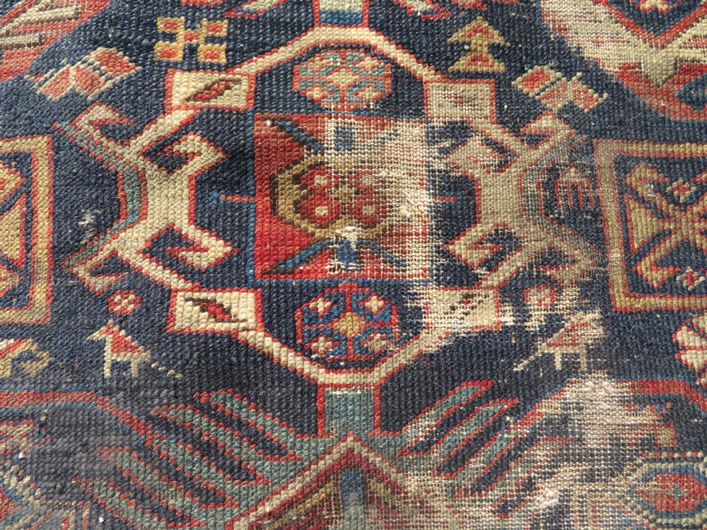 A rectangular hand-knotted antique Caucasian Daghestan rug featuring geometric medallions, intricate borders, and a distressed surface in red and navy blue tones. View III