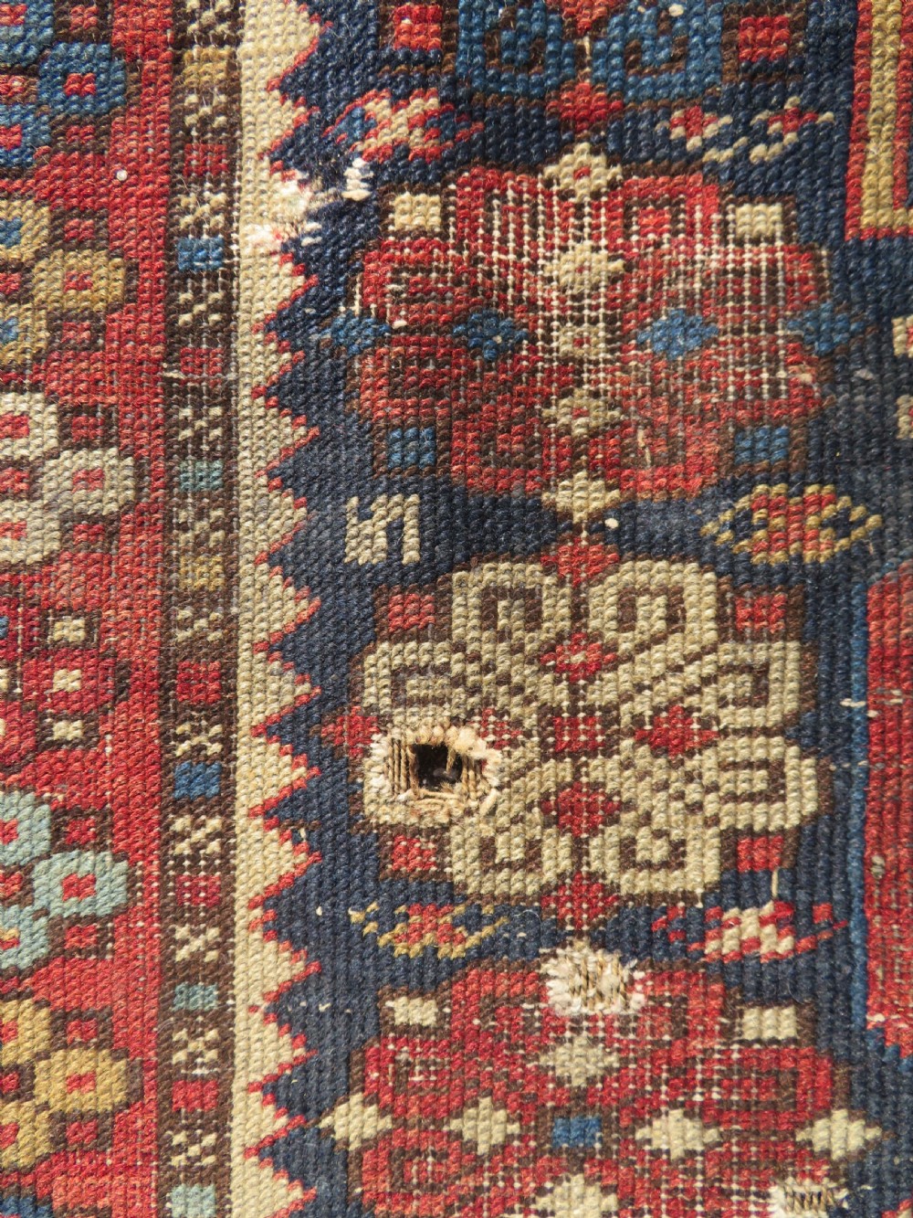 A rectangular hand-knotted antique Caucasian Daghestan rug featuring geometric medallions, intricate borders, and a distressed surface in red and navy blue tones. View IV