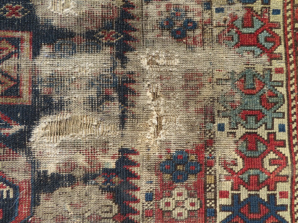 A rectangular hand-knotted antique Caucasian Daghestan rug featuring geometric medallions, intricate borders, and a distressed surface in red and navy blue tones. View V