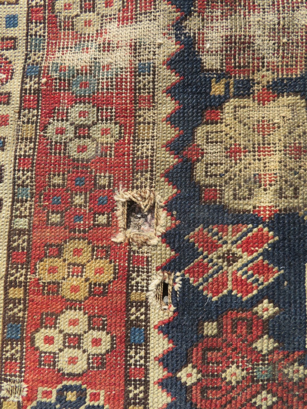 A rectangular hand-knotted antique Caucasian Daghestan rug featuring geometric medallions, intricate borders, and a distressed surface in red and navy blue tones. View VI