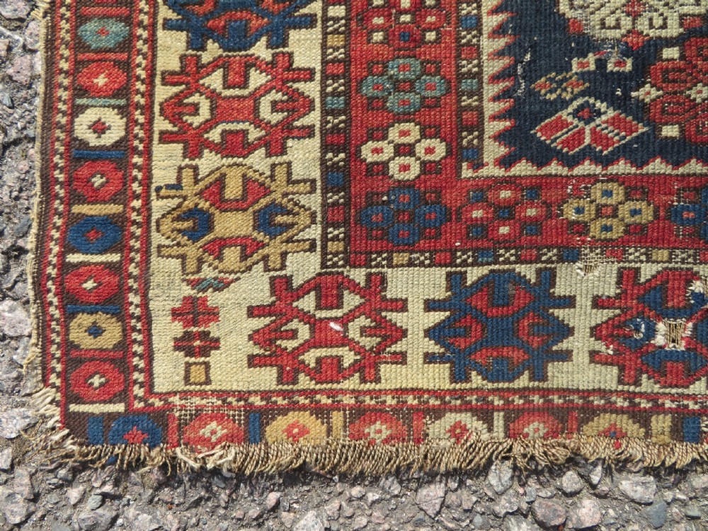 A rectangular hand-knotted antique Caucasian Daghestan rug featuring geometric medallions, intricate borders, and a distressed surface in red and navy blue tones. View VII