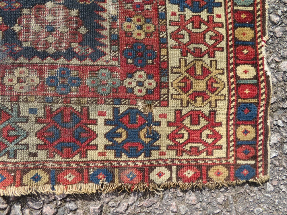 A rectangular hand-knotted antique Caucasian Daghestan rug featuring geometric medallions, intricate borders, and a distressed surface in red and navy blue tones. View VIII