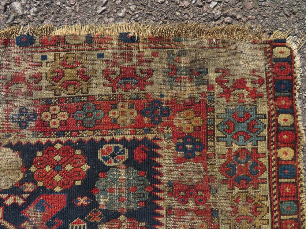 A rectangular hand-knotted antique Caucasian Daghestan rug featuring geometric medallions, intricate borders, and a distressed surface in red and navy blue tones. View X