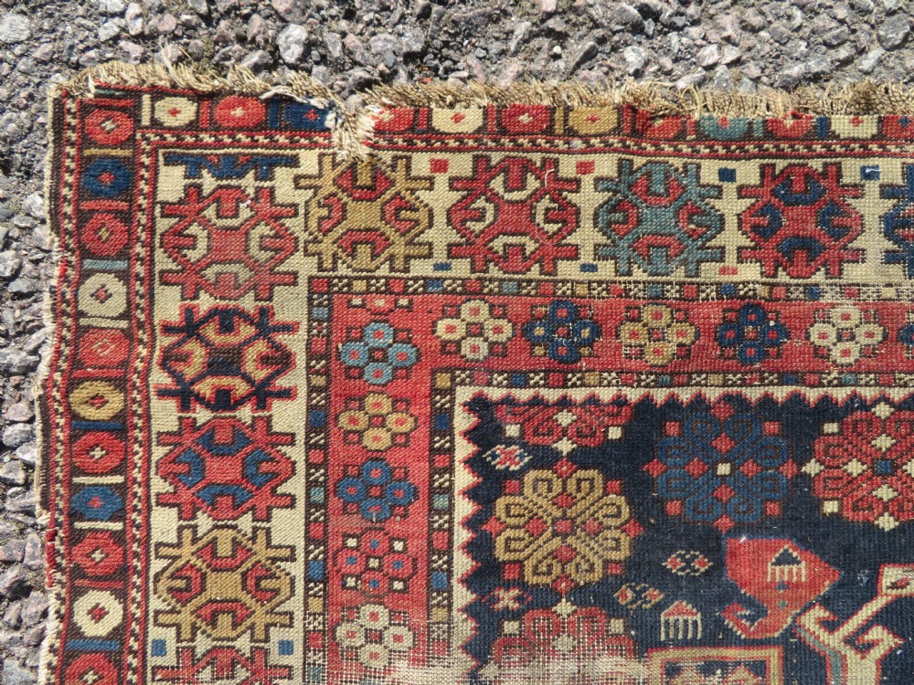 A rectangular hand-knotted antique Caucasian Daghestan rug featuring geometric medallions, intricate borders, and a distressed surface in red and navy blue tones. View XI