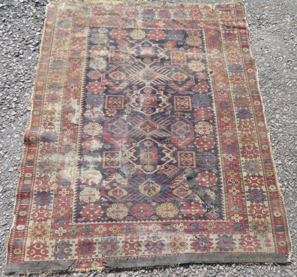 A rectangular hand-knotted antique Caucasian Daghestan rug featuring geometric medallions, intricate borders, and a distressed surface in red and navy blue tones. View XII