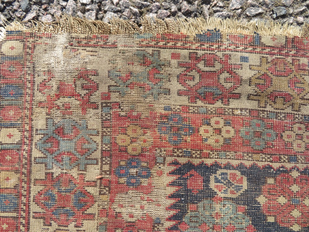 A rectangular hand-knotted antique Caucasian Daghestan rug featuring geometric medallions, intricate borders, and a distressed surface in red and navy blue tones. View XV