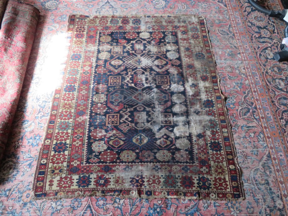 A rectangular hand-knotted antique Caucasian Daghestan rug featuring geometric medallions, intricate borders, and a distressed surface in red and navy blue tones. View XVII
