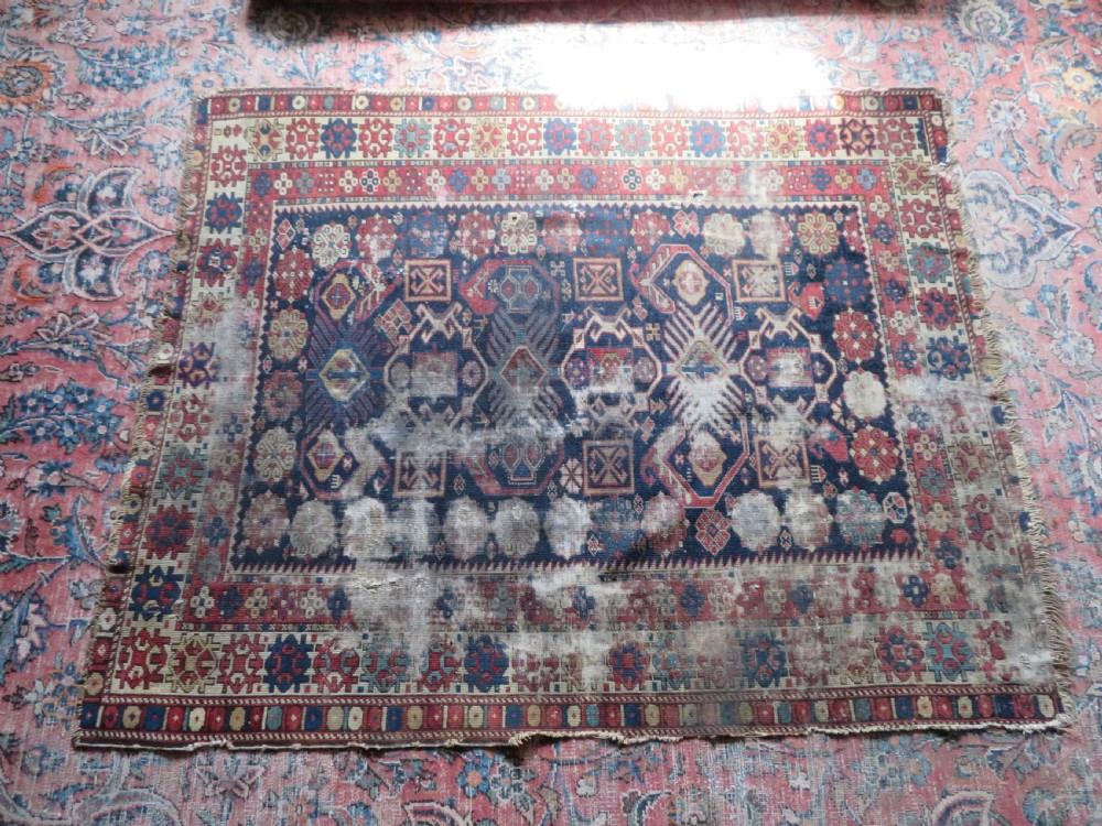 A rectangular hand-knotted antique Caucasian Daghestan rug featuring geometric medallions, intricate borders, and a distressed surface in red and navy blue tones. View XVIII