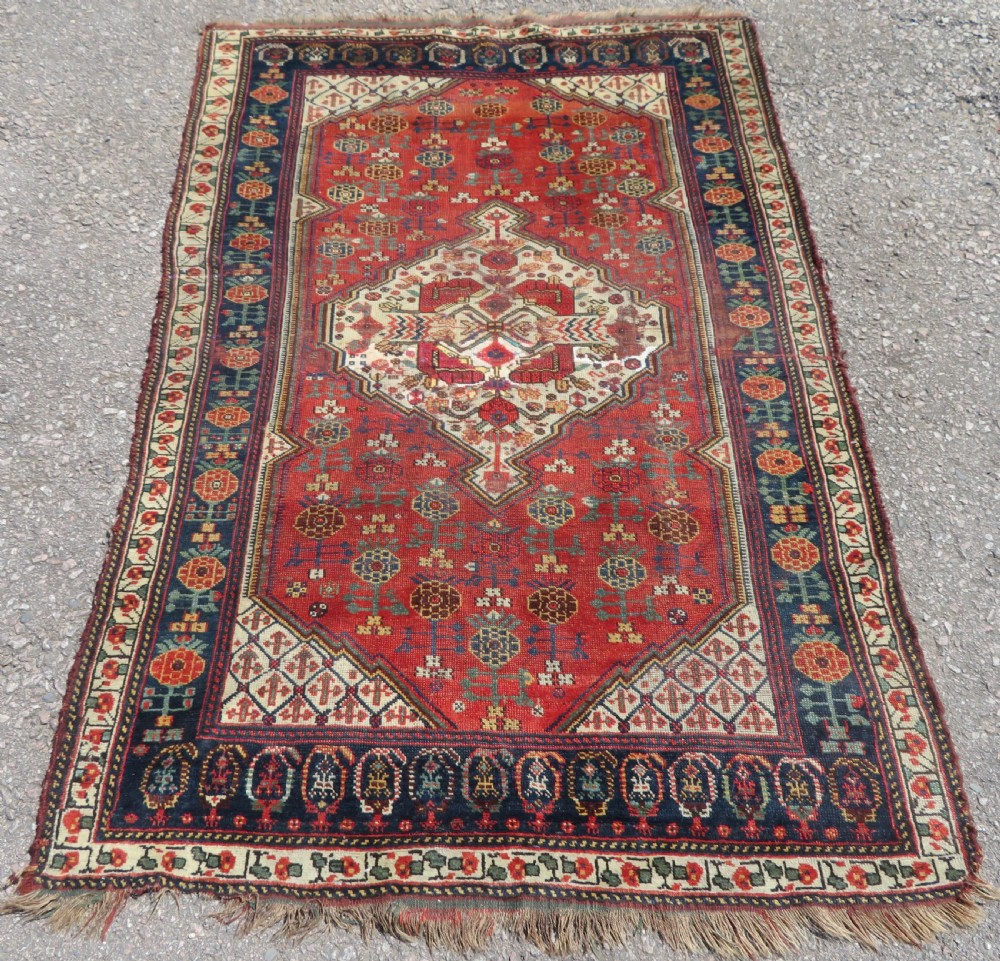 An antique hand-knotted Khamseh wool rug featuring a red central field with a geometric medallion framed by navy blue and cream borders.
