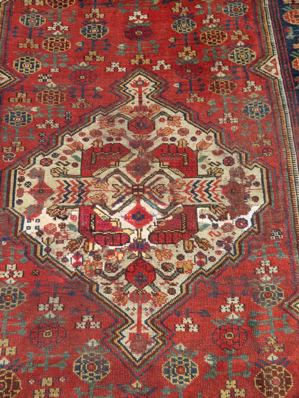 An antique hand-knotted Khamseh wool rug featuring a red central field with a geometric medallion framed by navy blue and cream borders. View III