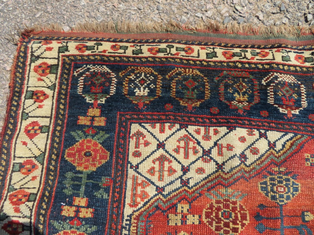 An antique hand-knotted Khamseh wool rug featuring a red central field with a geometric medallion framed by navy blue and cream borders. View X