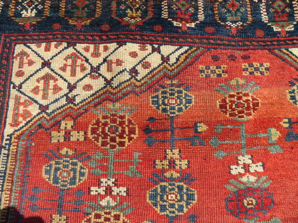 An antique hand-knotted Khamseh wool rug featuring a red central field with a geometric medallion framed by navy blue and cream borders. View XII