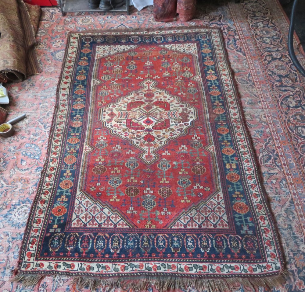 An antique hand-knotted Khamseh wool rug featuring a red central field with a geometric medallion framed by navy blue and cream borders. View XX