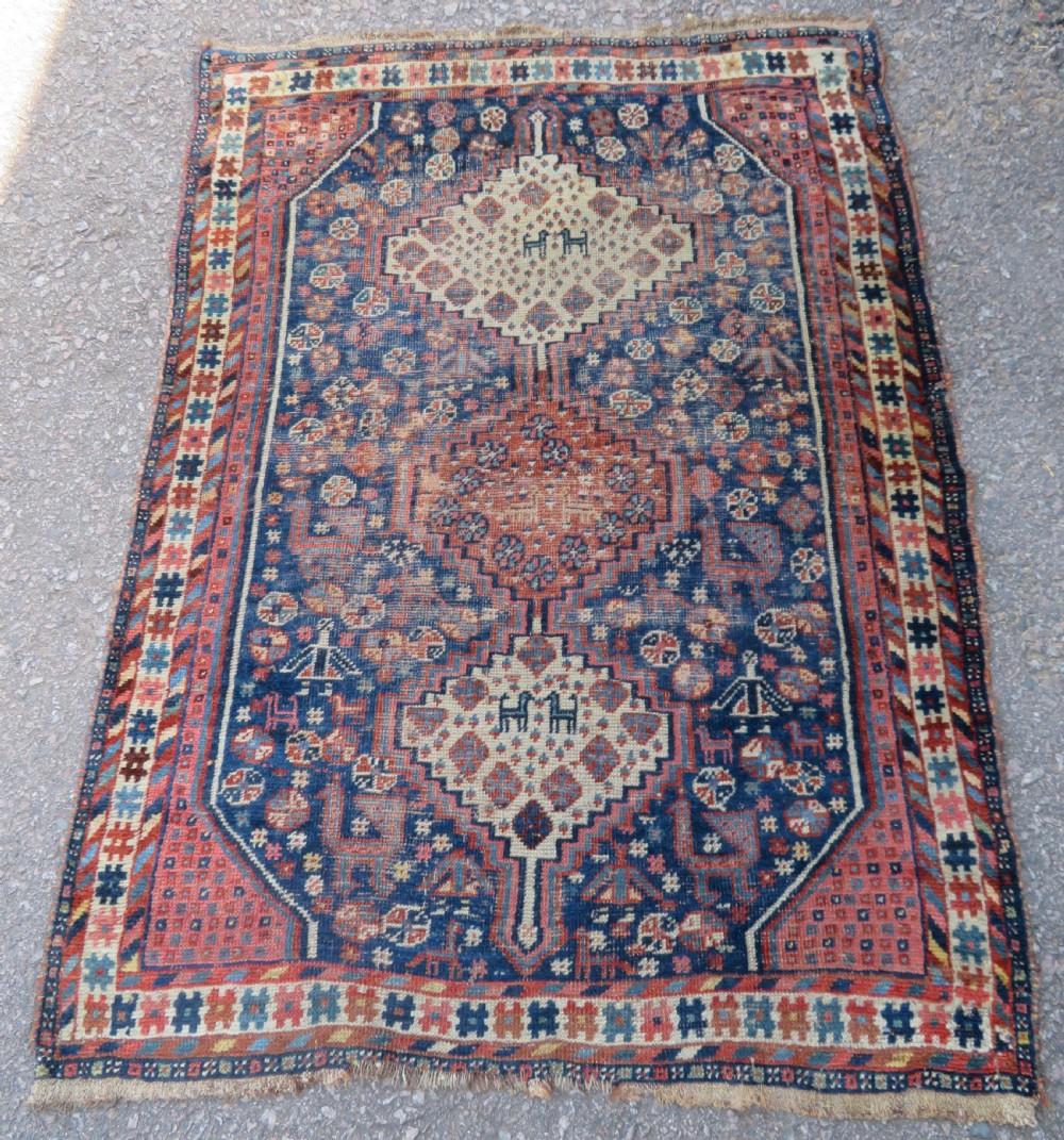 Distressed hand-knotted South Persian Khamseh tribal rug featuring nested diamond medallions, stylized animal motifs, and a multi-banded geometric border in navy blue, terracotta, and cream.