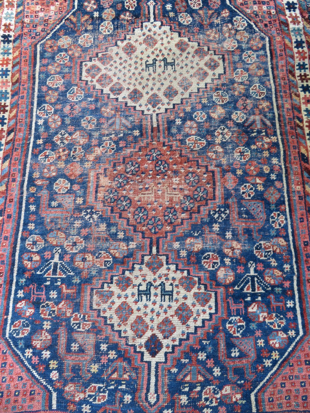 Distressed hand-knotted South Persian Khamseh tribal rug featuring nested diamond medallions, stylized animal motifs, and a multi-banded geometric border in navy blue, terracotta, and cream. View III