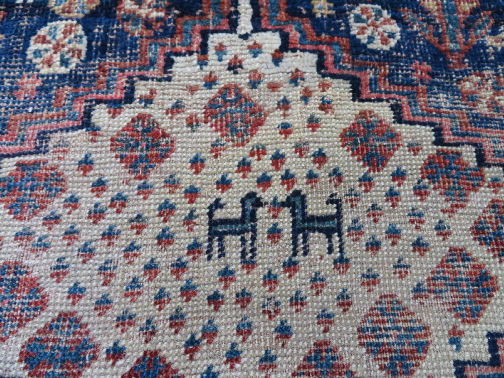 Distressed hand-knotted South Persian Khamseh tribal rug featuring nested diamond medallions, stylized animal motifs, and a multi-banded geometric border in navy blue, terracotta, and cream. View VII