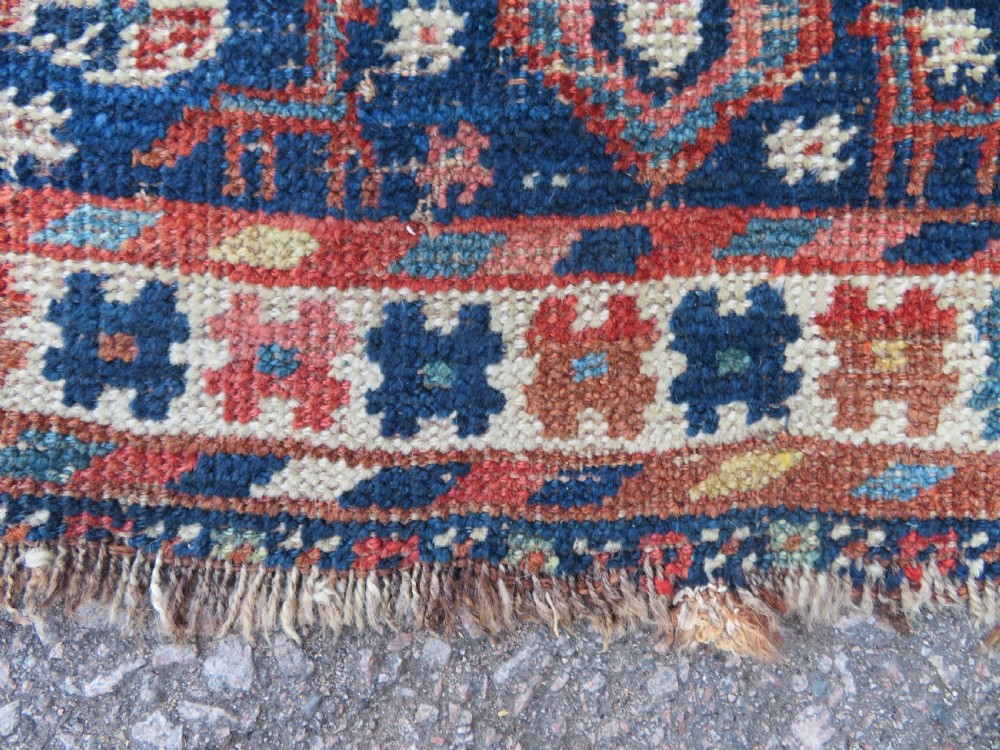 Distressed hand-knotted South Persian Khamseh tribal rug featuring nested diamond medallions, stylized animal motifs, and a multi-banded geometric border in navy blue, terracotta, and cream. View IX
