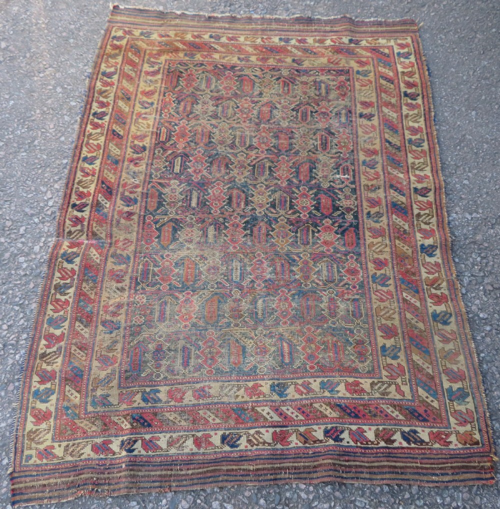 A rectangular hand-knotted wool rug displaying geometric repeating motifs in red, blue, and tan with visible wear and distressed surface texture.