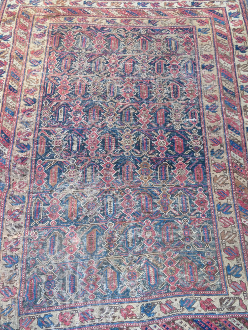 A rectangular hand-knotted wool rug displaying geometric repeating motifs in red, blue, and tan with visible wear and distressed surface texture. View II