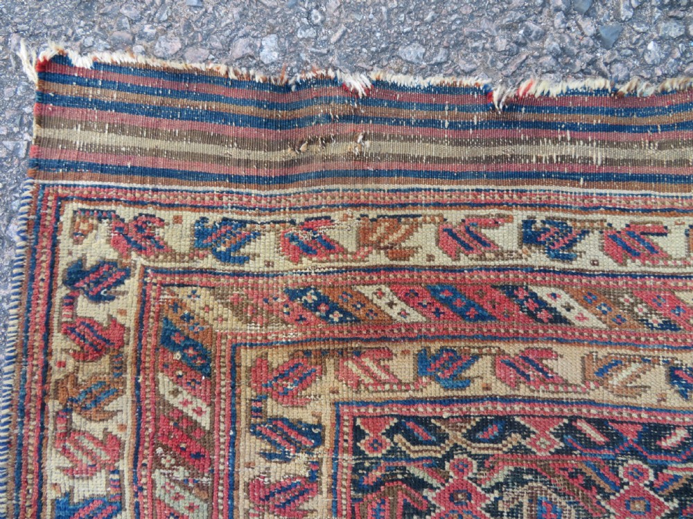 A rectangular hand-knotted wool rug displaying geometric repeating motifs in red, blue, and tan with visible wear and distressed surface texture. View III