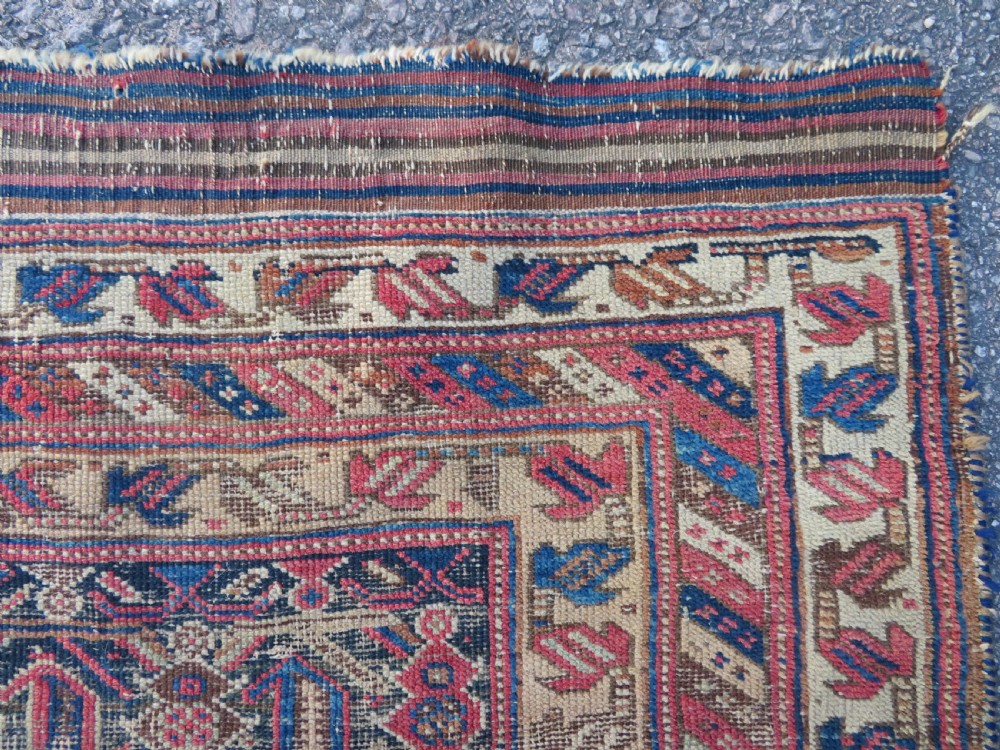 A rectangular hand-knotted wool rug displaying geometric repeating motifs in red, blue, and tan with visible wear and distressed surface texture. View IV