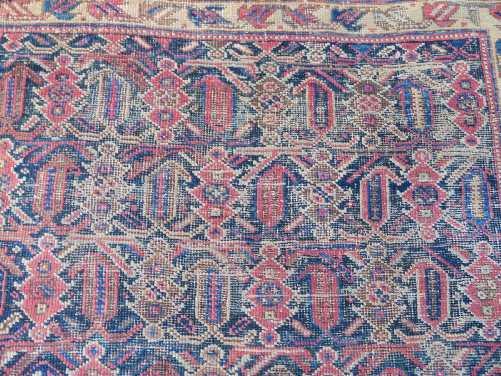 A rectangular hand-knotted wool rug displaying geometric repeating motifs in red, blue, and tan with visible wear and distressed surface texture. View V