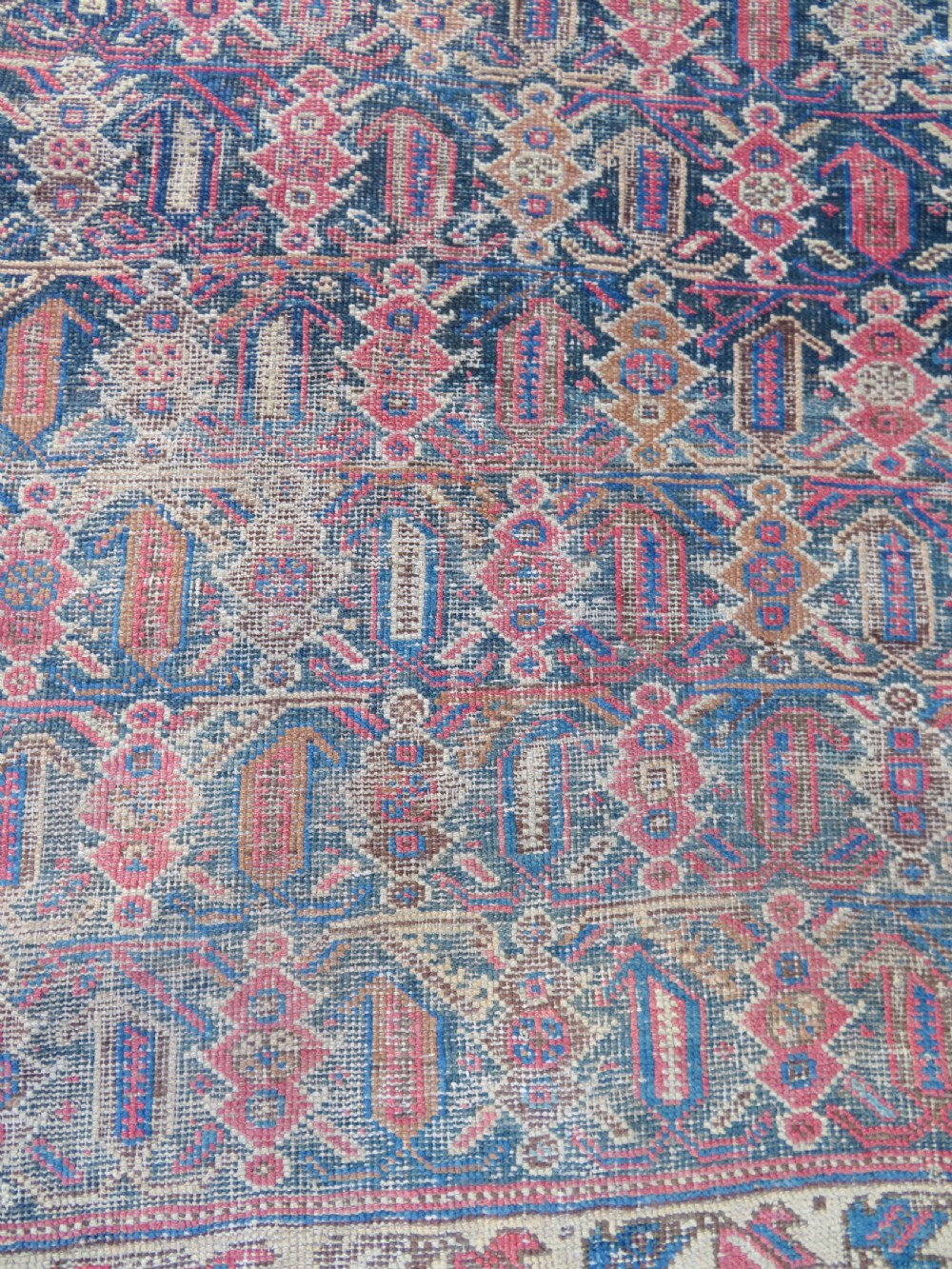 A rectangular hand-knotted wool rug displaying geometric repeating motifs in red, blue, and tan with visible wear and distressed surface texture. View VI