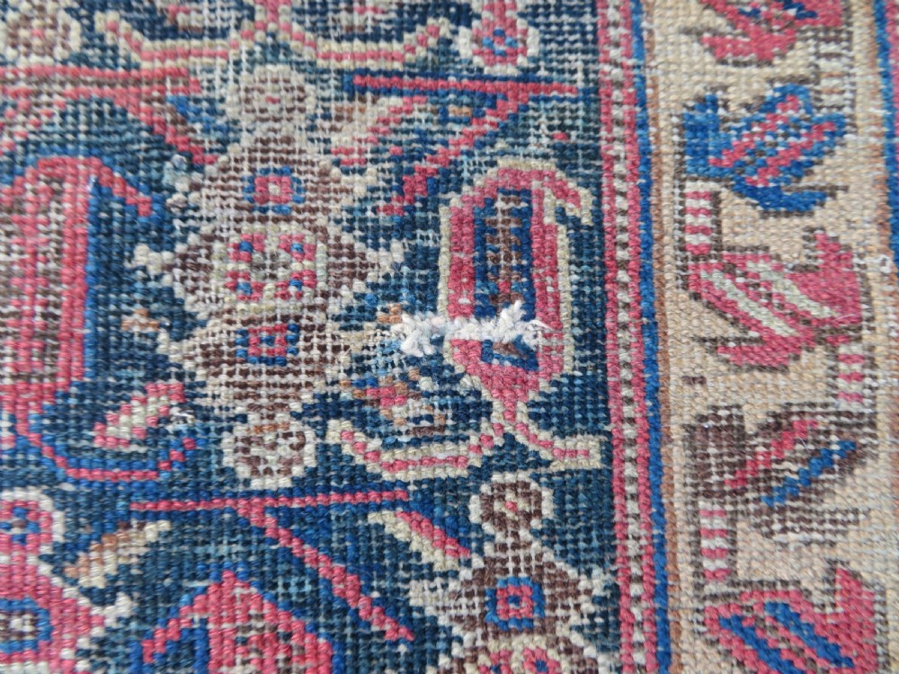 A rectangular hand-knotted wool rug displaying geometric repeating motifs in red, blue, and tan with visible wear and distressed surface texture. View VII