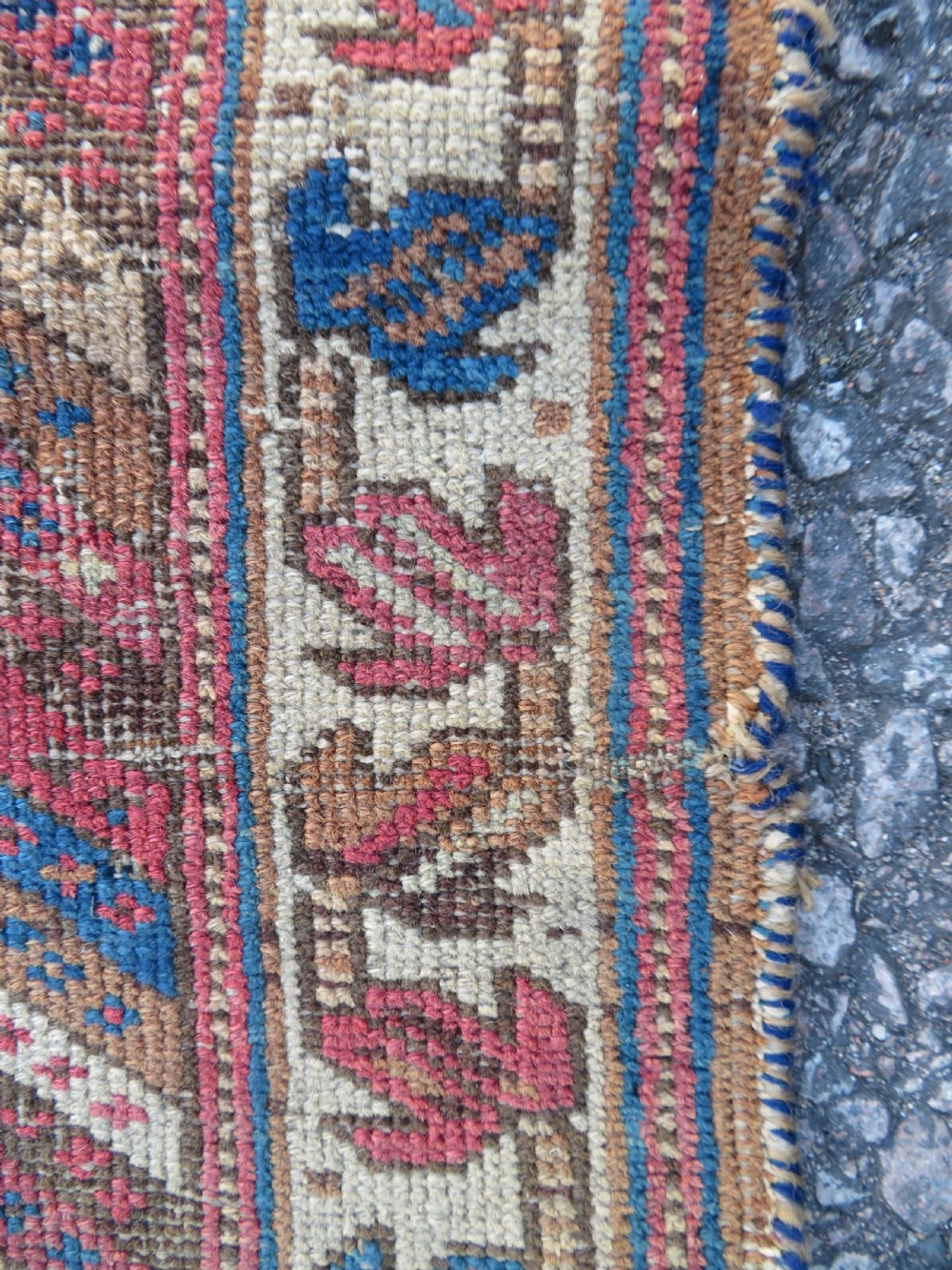 A rectangular hand-knotted wool rug displaying geometric repeating motifs in red, blue, and tan with visible wear and distressed surface texture. View VIII