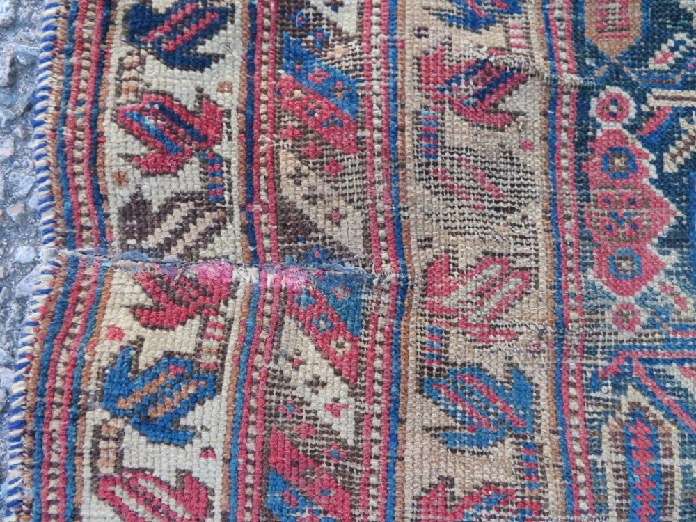 A rectangular hand-knotted wool rug displaying geometric repeating motifs in red, blue, and tan with visible wear and distressed surface texture. View IX