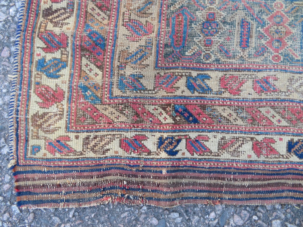 A rectangular hand-knotted wool rug displaying geometric repeating motifs in red, blue, and tan with visible wear and distressed surface texture. View X