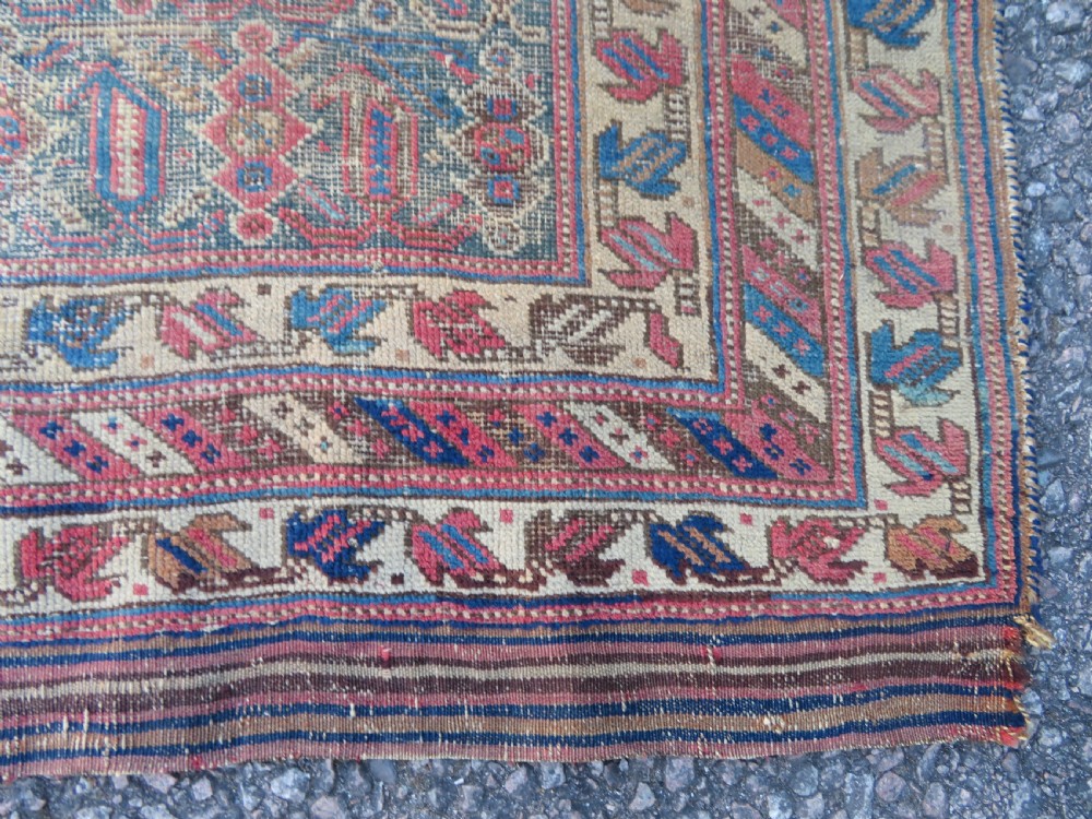 A rectangular hand-knotted wool rug displaying geometric repeating motifs in red, blue, and tan with visible wear and distressed surface texture. View XI