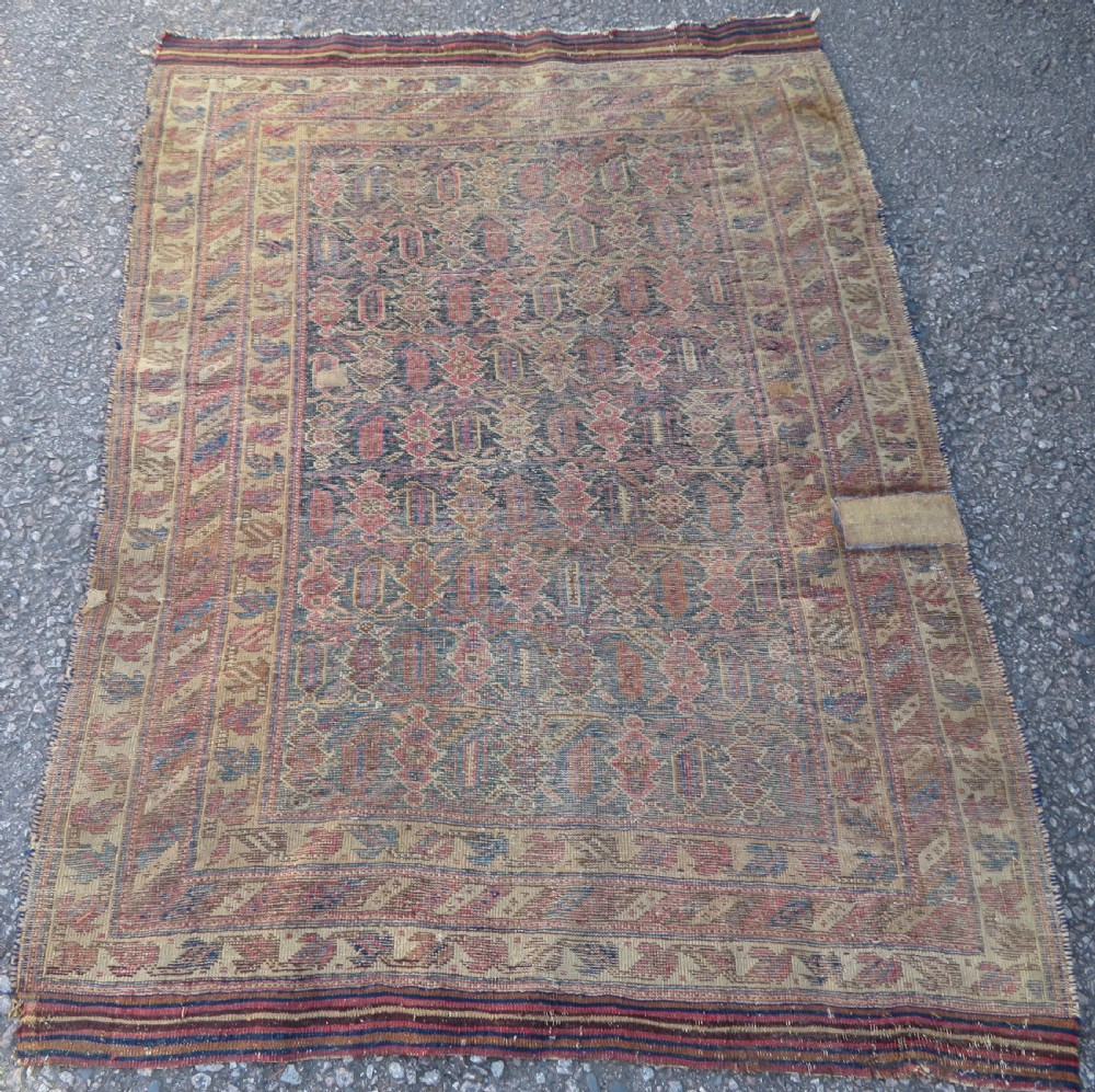 A rectangular hand-knotted wool rug displaying geometric repeating motifs in red, blue, and tan with visible wear and distressed surface texture. View XII
