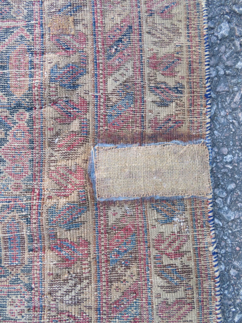 A rectangular hand-knotted wool rug displaying geometric repeating motifs in red, blue, and tan with visible wear and distressed surface texture. View XIII
