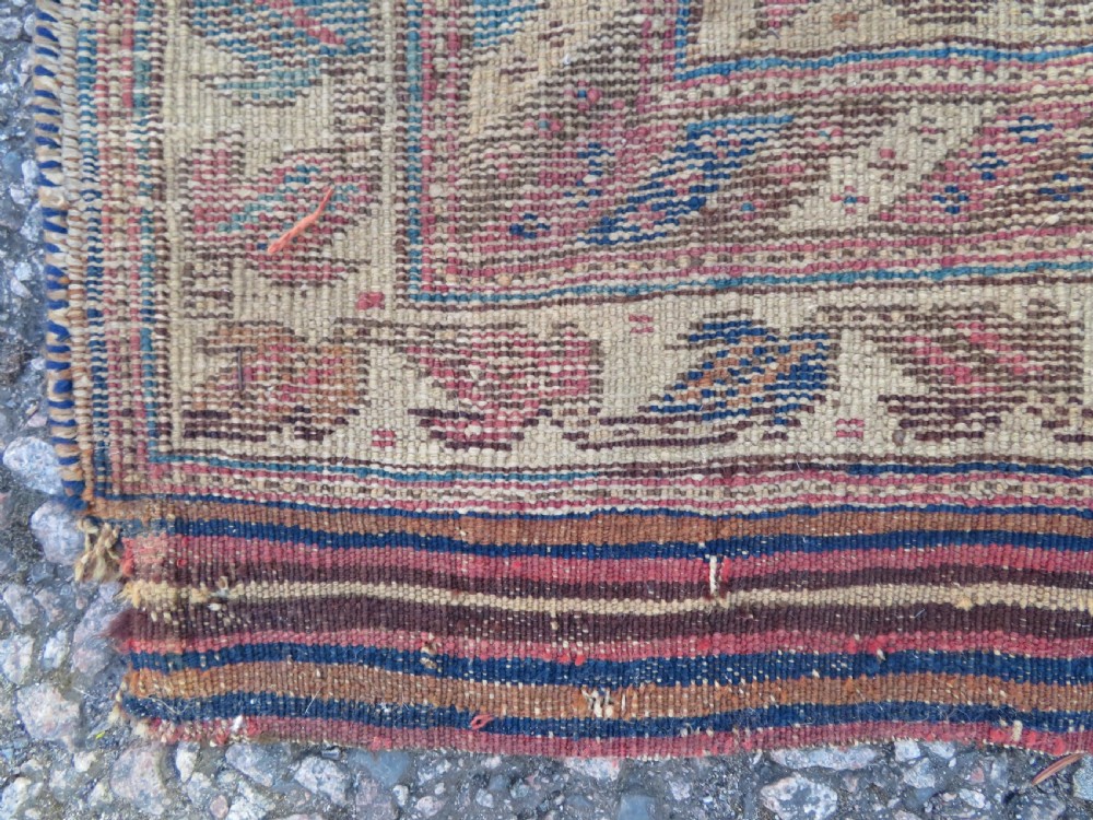 A rectangular hand-knotted wool rug displaying geometric repeating motifs in red, blue, and tan with visible wear and distressed surface texture. View XV