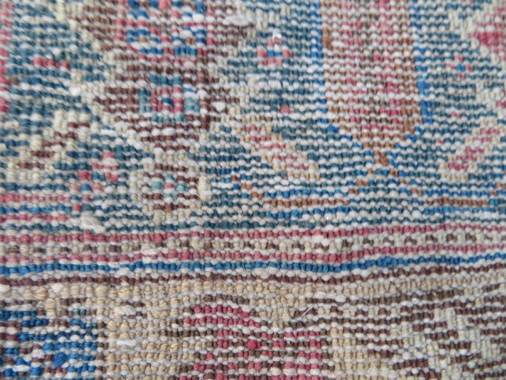 A rectangular hand-knotted wool rug displaying geometric repeating motifs in red, blue, and tan with visible wear and distressed surface texture. View XVI