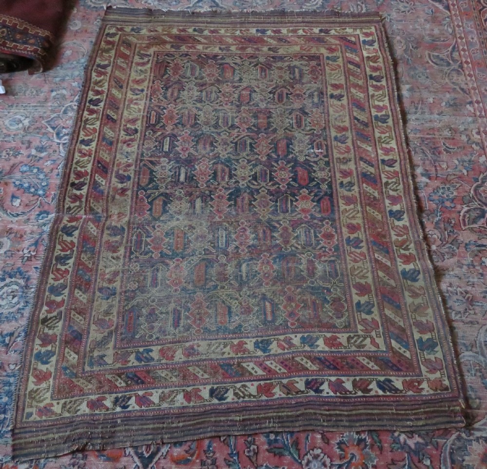 A rectangular hand-knotted wool rug displaying geometric repeating motifs in red, blue, and tan with visible wear and distressed surface texture. View XVII