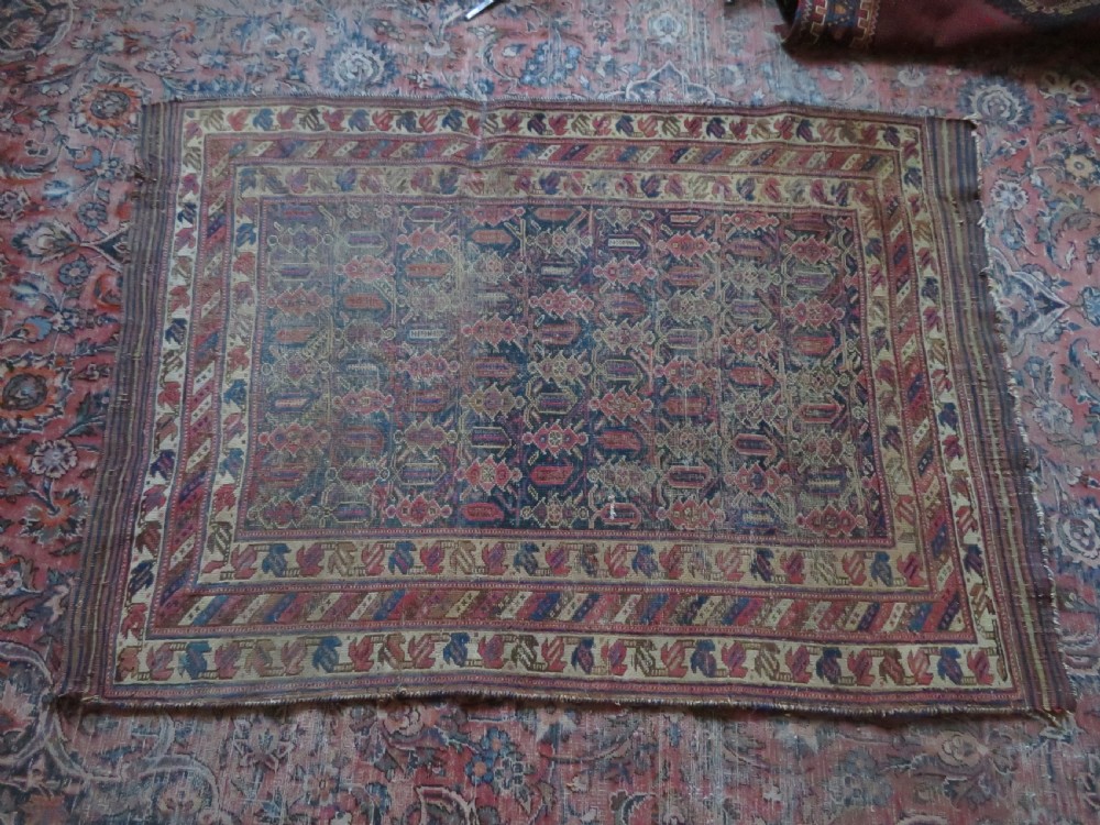 A rectangular hand-knotted wool rug displaying geometric repeating motifs in red, blue, and tan with visible wear and distressed surface texture. View XVIII