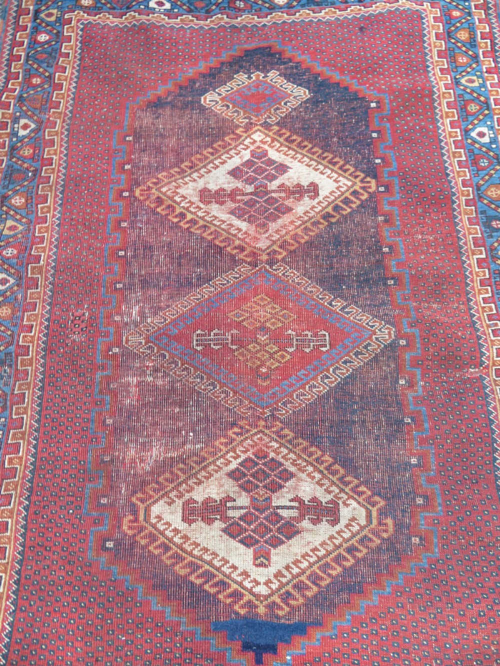 A rectangular hand-knotted wool rug featuring three diamond medallions on a dark central field surrounded by a patterned terracotta red background and geometric borders. View II