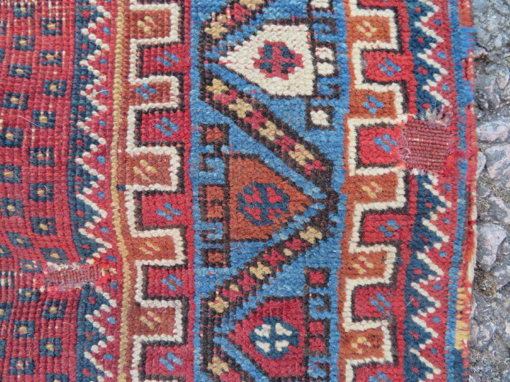A rectangular hand-knotted wool rug featuring three diamond medallions on a dark central field surrounded by a patterned terracotta red background and geometric borders. View IV